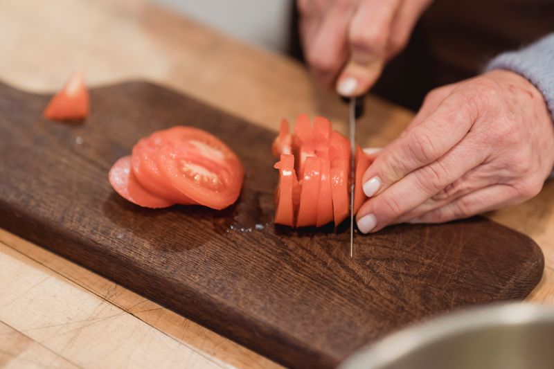Crop anonymous female chef with manicured hands slicing organic ripe tomato on wooden board while cooking