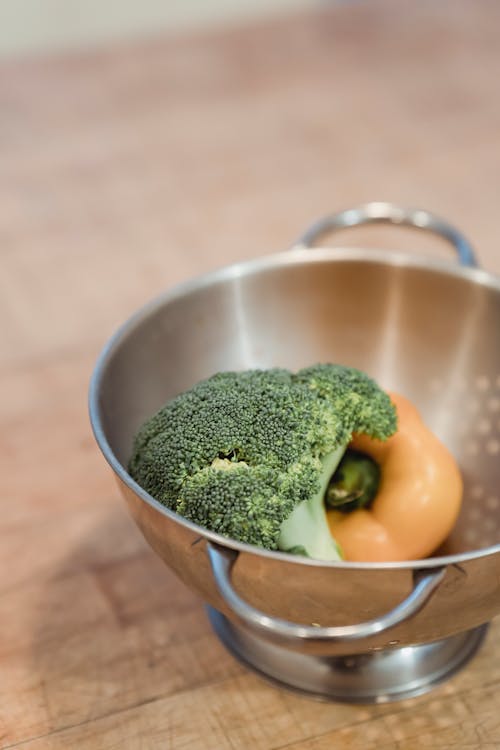 Raw ripe broccoli and yellow capsicum placed in steel colander on wooden table in kitchen