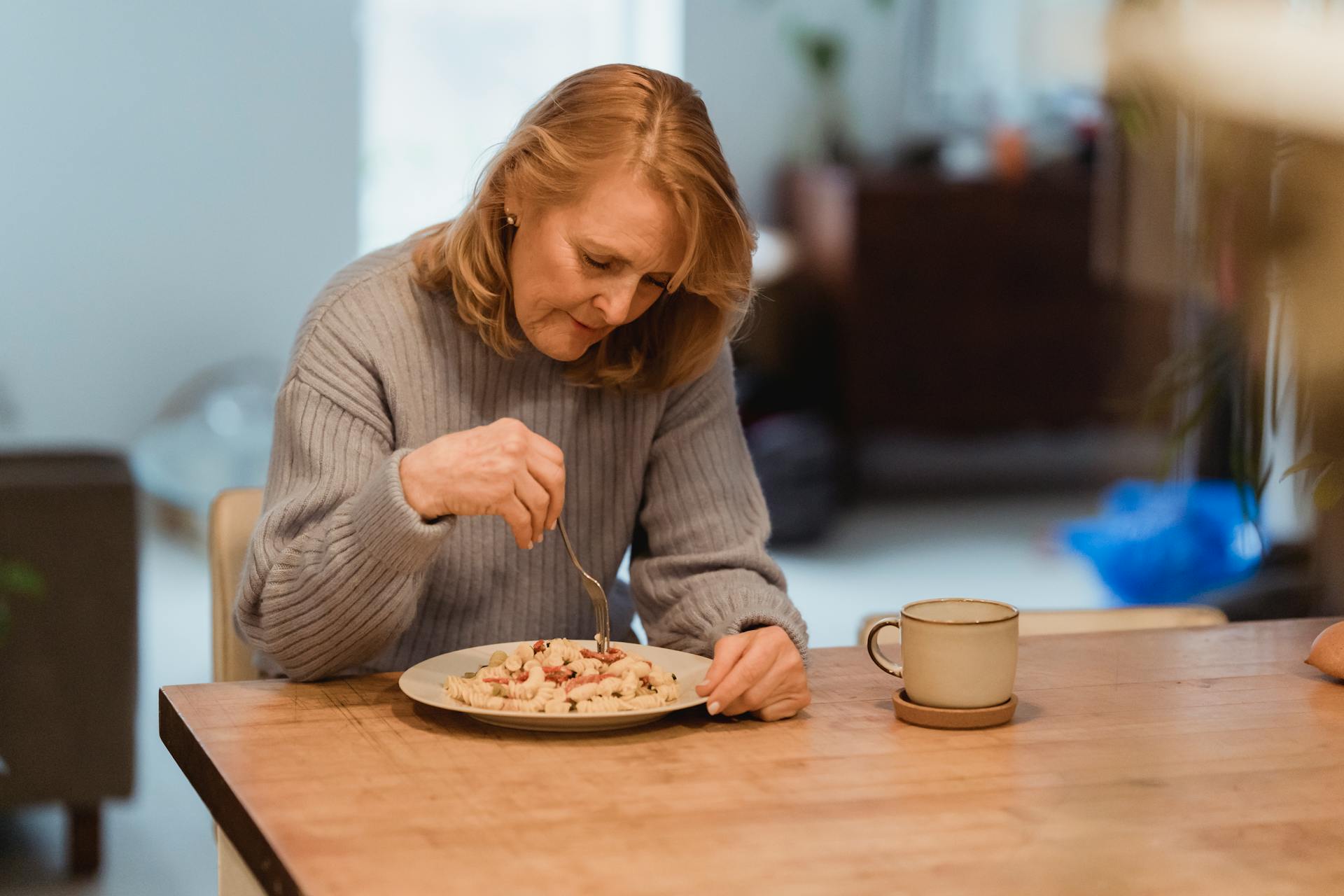 Emotionless senior female wearing blue sweater enjoying yummy vegetable pasta while sitting at table in living room