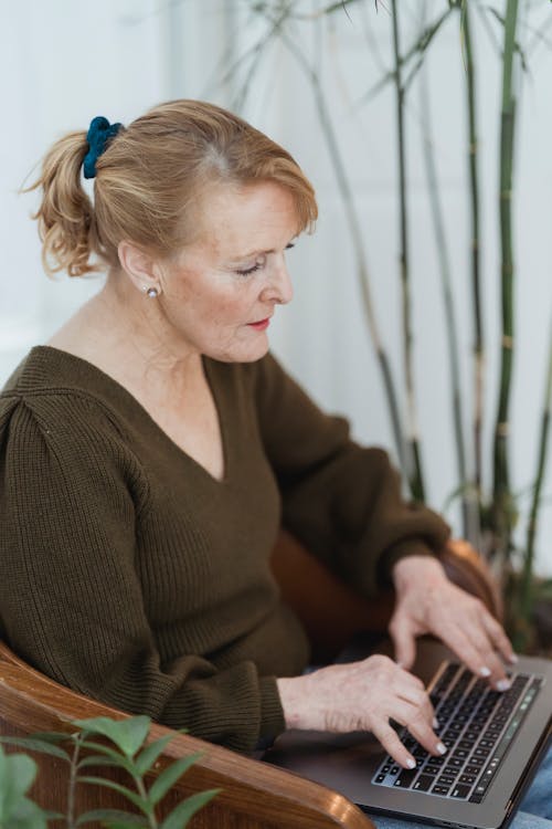 Free Serious mature female in casual outfit typing on modern netbook keyboard while sitting on comfy armchair in light living room Stock Photo