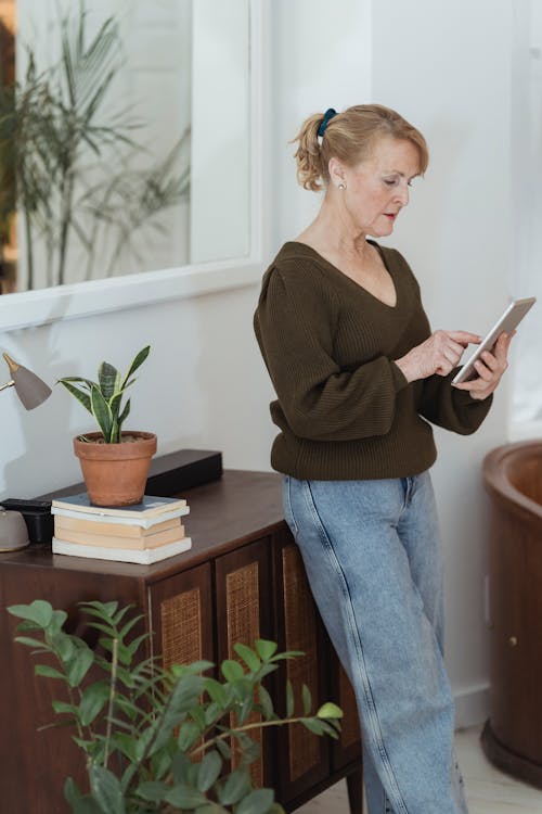 Focused mature female in jeans and sweater browsing modern tablet and standing in modern light living room