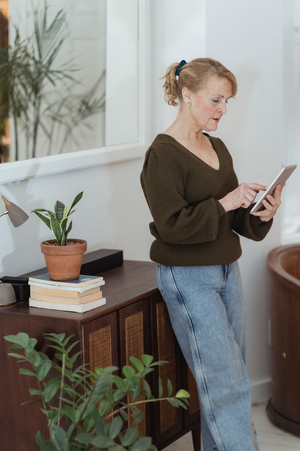 A mature woman in a green sweater focuses on a tablet in her warmly lit living room at home