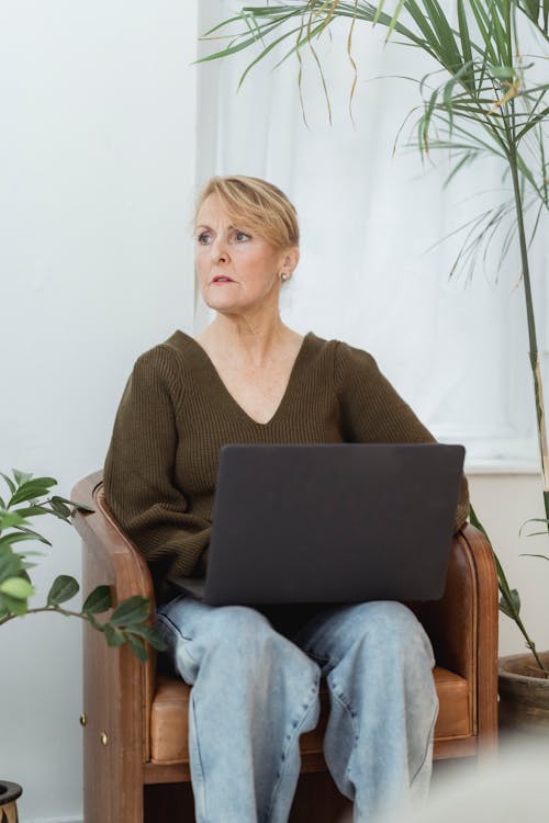 Pensive mature woman working on laptop on cozy armchair