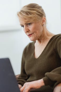 Focused mature female in casual sweater surfing internet on modern netbook in light room