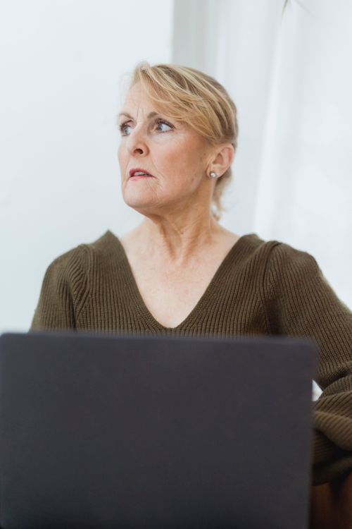 Free Positive mature female wearing casual sweater browsing modern netbook in light room and looking away thoughtfully Stock Photo