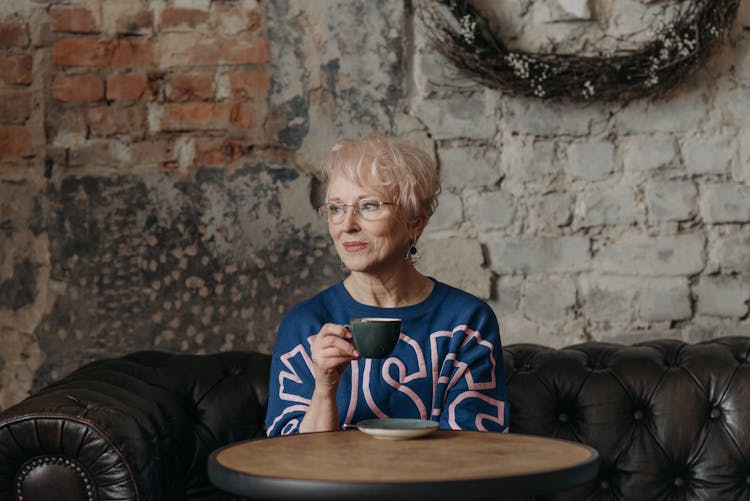 Blonde Elderly Woman Sitting On Black Sofa Having Coffee