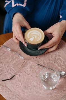 A top view of hands holding a latte with intricate latte art on a crocheted tablecloth.