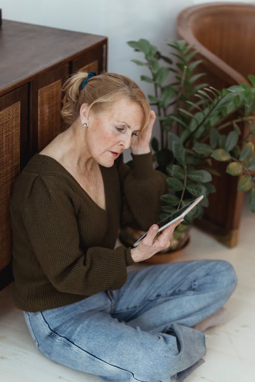 Free Pensive mature female touching hair while sitting with crossed legs on floor with cellphone at home Stock Photo