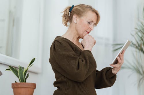 Side view of thoughtful elderly female in sweater with cellphone touching chin against potted plant in house room