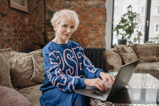 Elderly woman with white hair using a laptop indoors, enjoying a warm, stylish workspace.