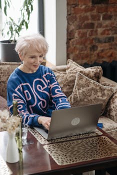 Elderly woman typing on laptop, seated comfortably at home, smiling.