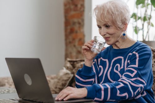 Free Elderly woman sipping water while working on a laptop in a cozy indoor setting. Stock Photo