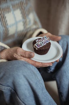 Chocolate muffin on a plate with 'Happy Birthday' decoration held by a person.