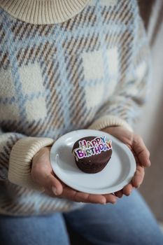 Crop anonymous female in sweater with checkered ornament holding plate with tasty chocolate dessert with decorative title on birthday