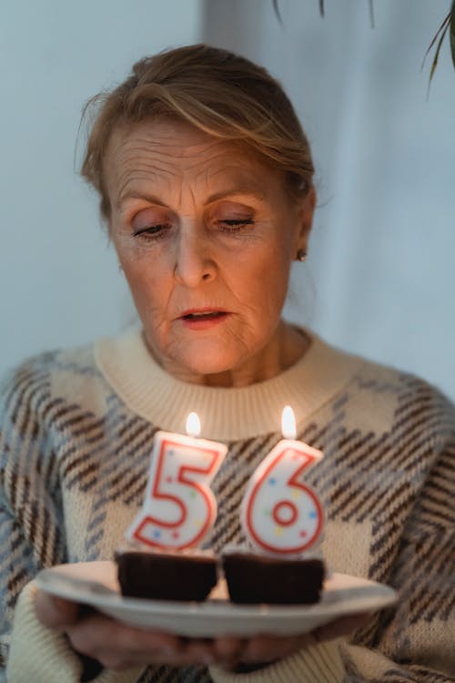 Mature female in cozy sweater holding plate with delicious birthday cakes and burning candles in shape of numbers
