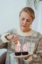 Senior female in casual sweater lightning candles in shape of numbers placed on birthday chocolate cupcakes