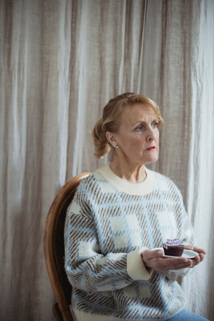 Pensive Mature Woman Holding Birthday Chocolate Cupcake In Light Room