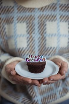 Crop anonymous female in warm sweater demonstrating plate with chocolate cupcake with decorative inscription Happy Birthday