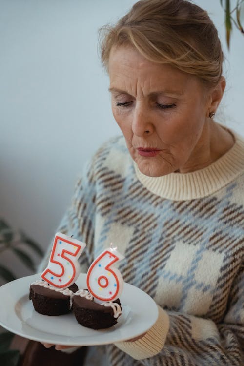 Crop senior female wearing sweater blowing candles on delicious cupcakes during birthday celebration