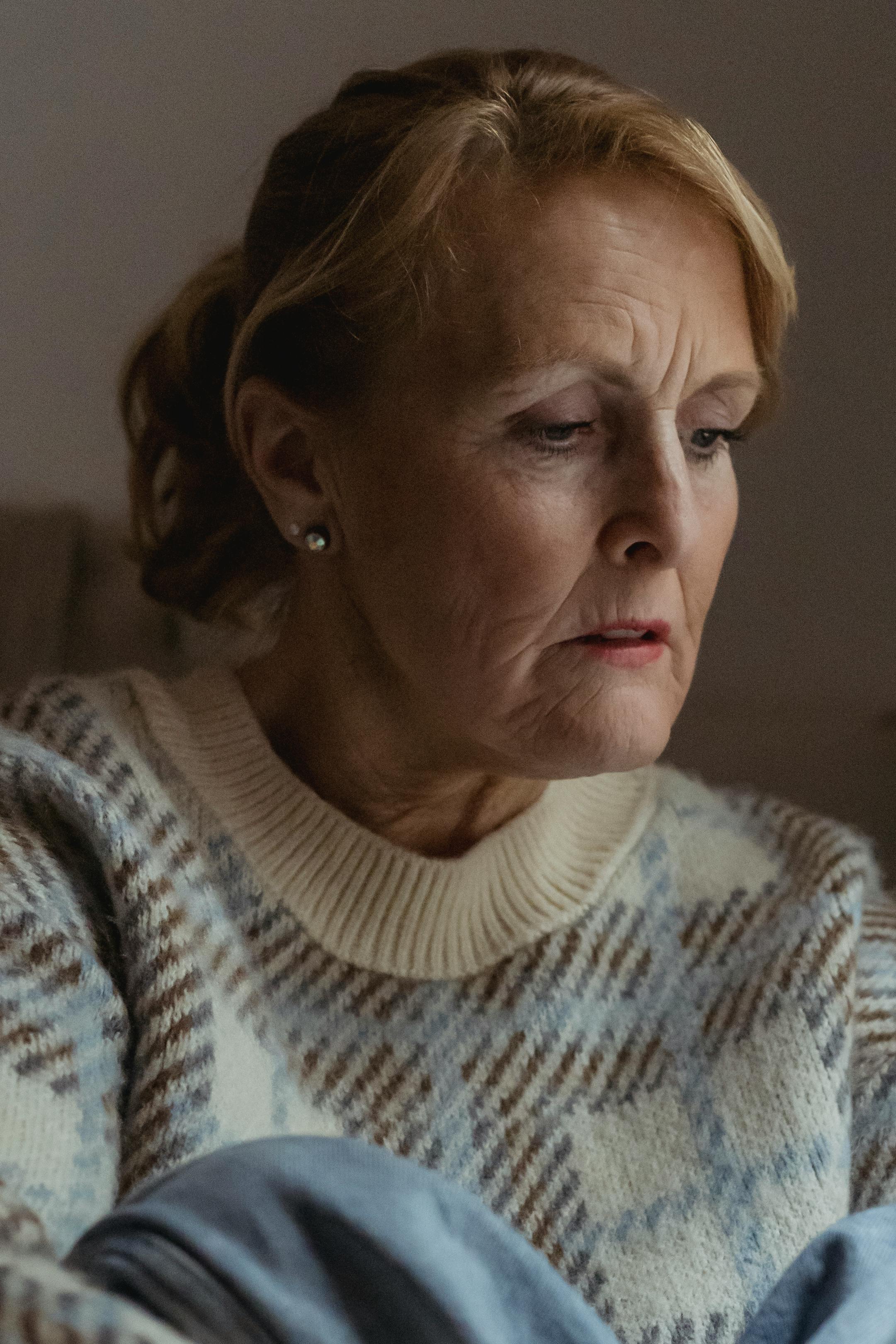 Preoccupied concerned senior female wearing casual clothes embracing knees and looking down in deep thoughts