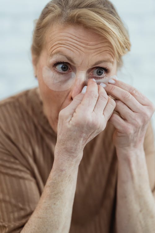 Crop focused mature female in casual wear applying anti aging under eye patches in light room