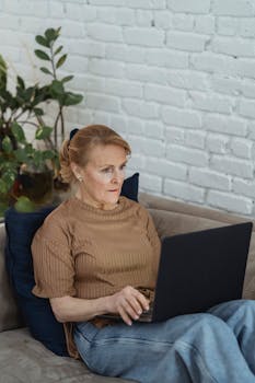 Concentrated senior female wearing casual outfit and hydrating eye patches working on modern netbook and lying on cozy sofa in modern living room