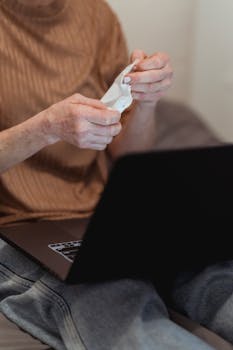 Senior woman using tissue while sitting with a laptop indoors, focusing on comfort and modern lifestyle.