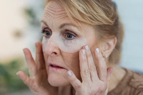 Free Crop unemotional mature female applying anti aging moisturizing eye patches and looking away in light room Stock Photo