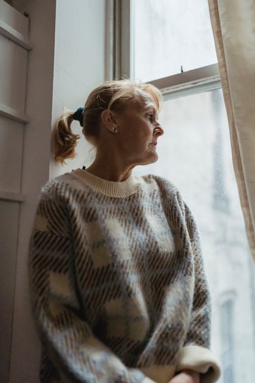 Contemplative senior female wearing warm sweater sitting on windowsill and looking out window in contemplation