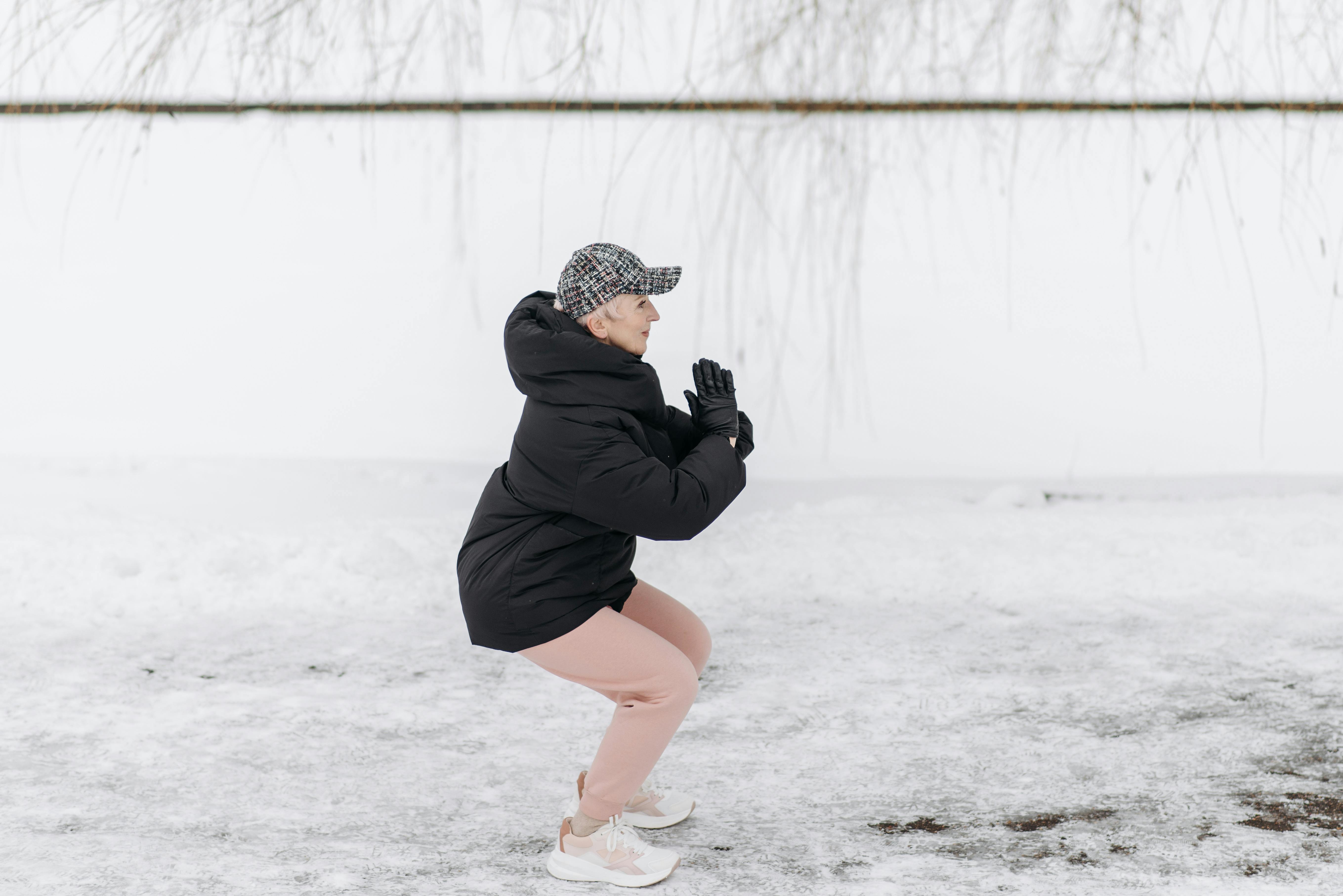 Senior woman doing squats outside in winter clothing on snowy ground.