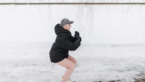 Senior woman doing squats outside in winter clothing on snowy ground.