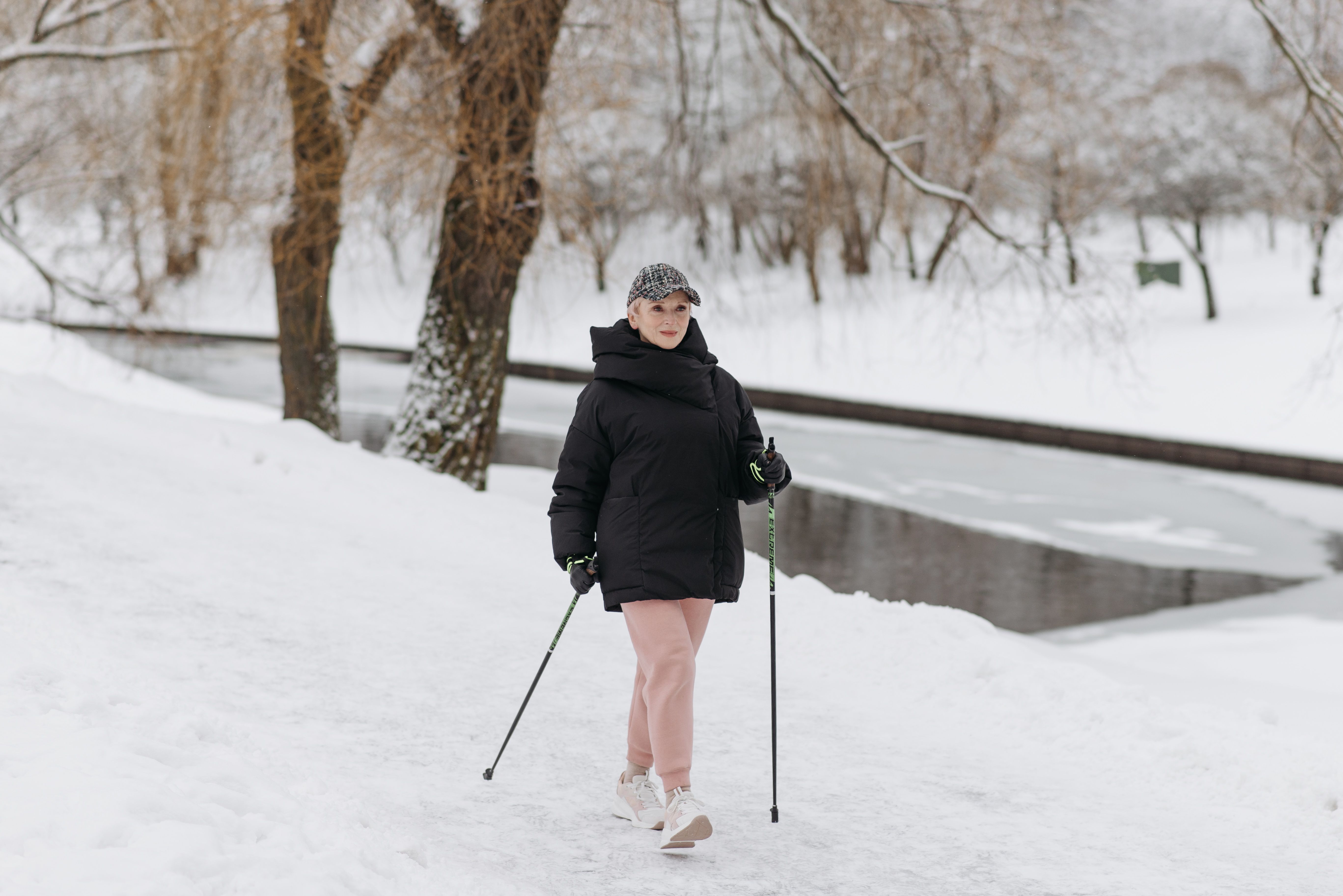 Caucasian elderly woman hiking in winter with trekking poles by a snowy riverbank.