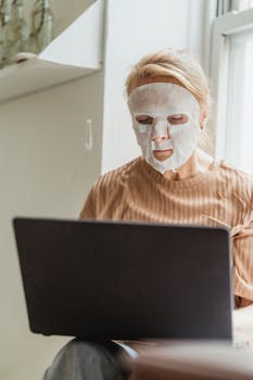 Senior woman multitasking with a cosmetic facial mask while working on a laptop indoors.
