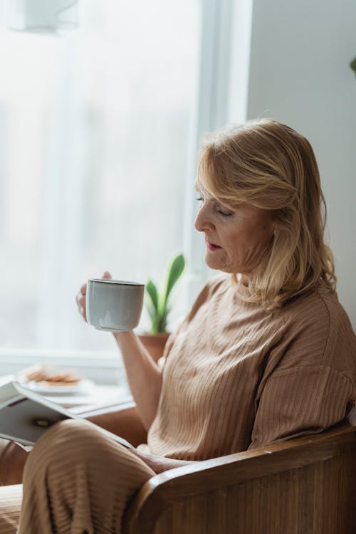 Free An Elderly Woman Sitting on a Sofa Chair Reading Book while Drinking a Cup of Coffee Stock Photo