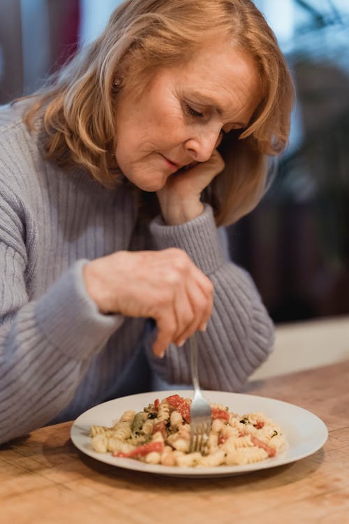 Free Unhappy mature female with fork sitting at table with plate of delicious pasta while having dinner in kitchen on blurred background Stock Photo