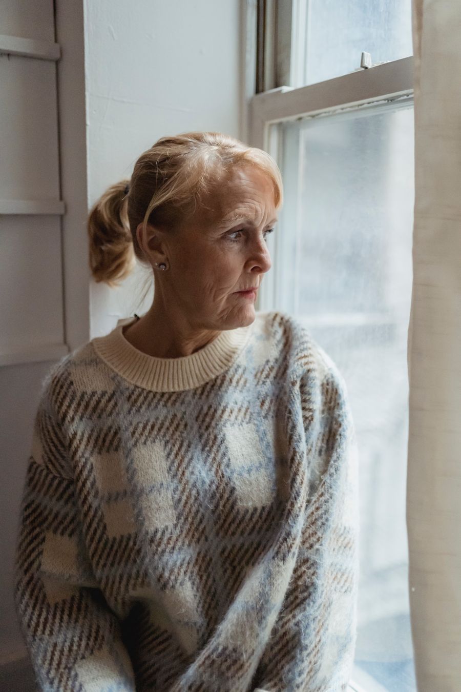 A calm woman in a warm sweater sitting by a window, thinking something over in soft daylight.
