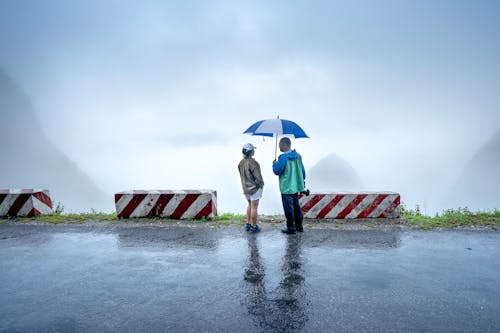 Man and Woman Standing Under an Umbrella while Standing on a Wet Road Near Concrete Barriers