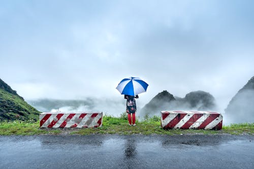 Back View Shot of a Person Holding an Umbrella while Standing on the Side of the Road Beside the Concrete Barriers