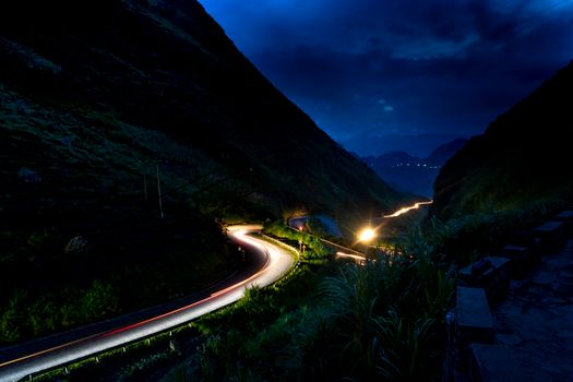 Serene nighttime view of a mountain road illuminated by car lights under a dramatic sky.