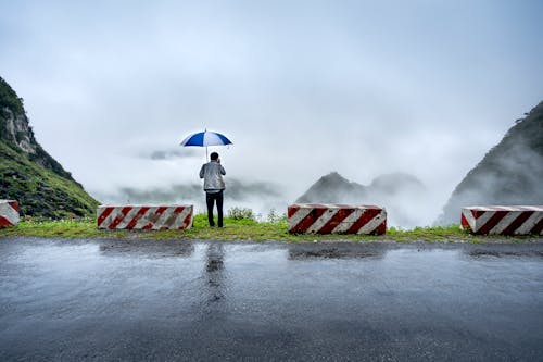 A Man Holding an Open Umbrella Standing on the Side of the Road while Looking at the Scenery