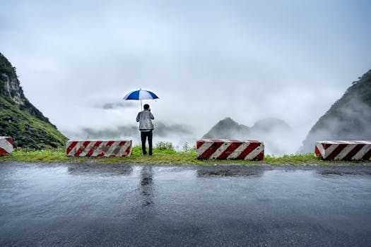 A man with an umbrella stands on a rainy road, looking at a foggy mountain landscape.