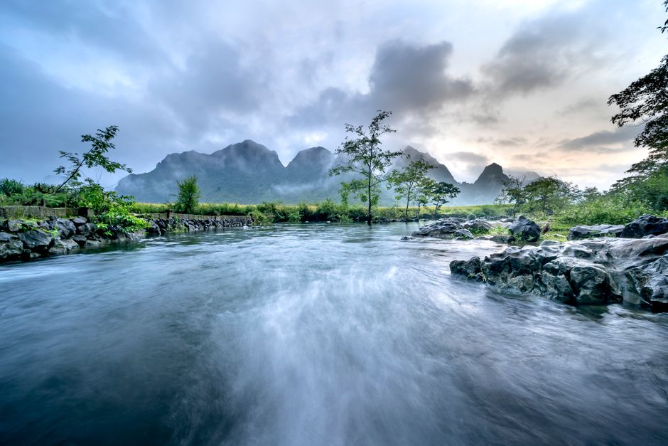 Fotomural Un río tranquilo fluye a través de un valle exuberante con montañas brumosas al fondo.