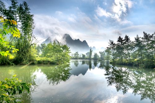 Free Peaceful river scene with mountain reflections and lush greenery under a bright sky. Stock Photo