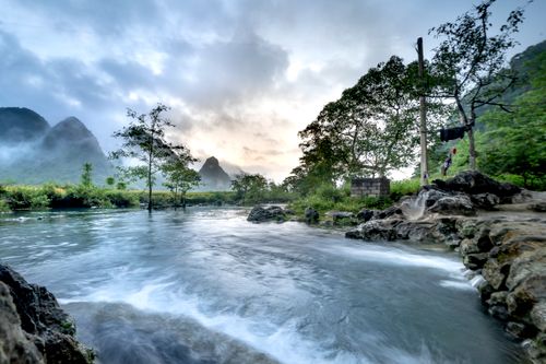 Free Serene river flowing amidst lush greenery and misty mountains at sunrise. Stock Photo