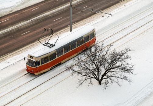 Aerial view of a red tram moving through snowy tracks with a leafless tree nearby, signaling winter.