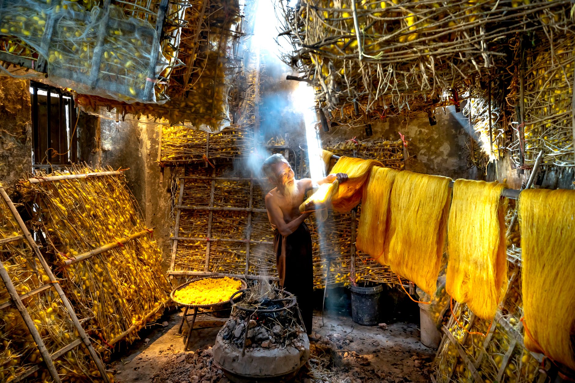 Elderly man working with silk threads in traditional workshop with sunlight streaming in.