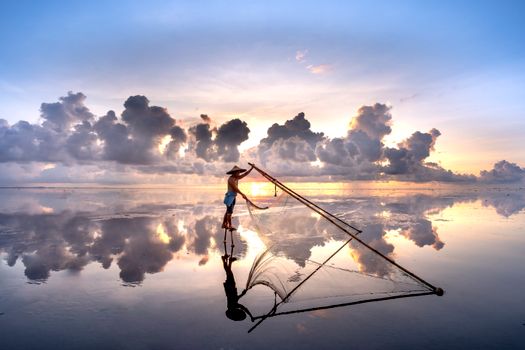 A fisherman in a conical hat casting a net over calm waters during sunset.