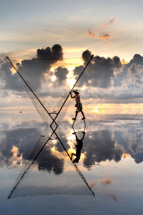 Free Silhouette of a fisherman casting net at sunset, reflecting on water surface. Stock Photo