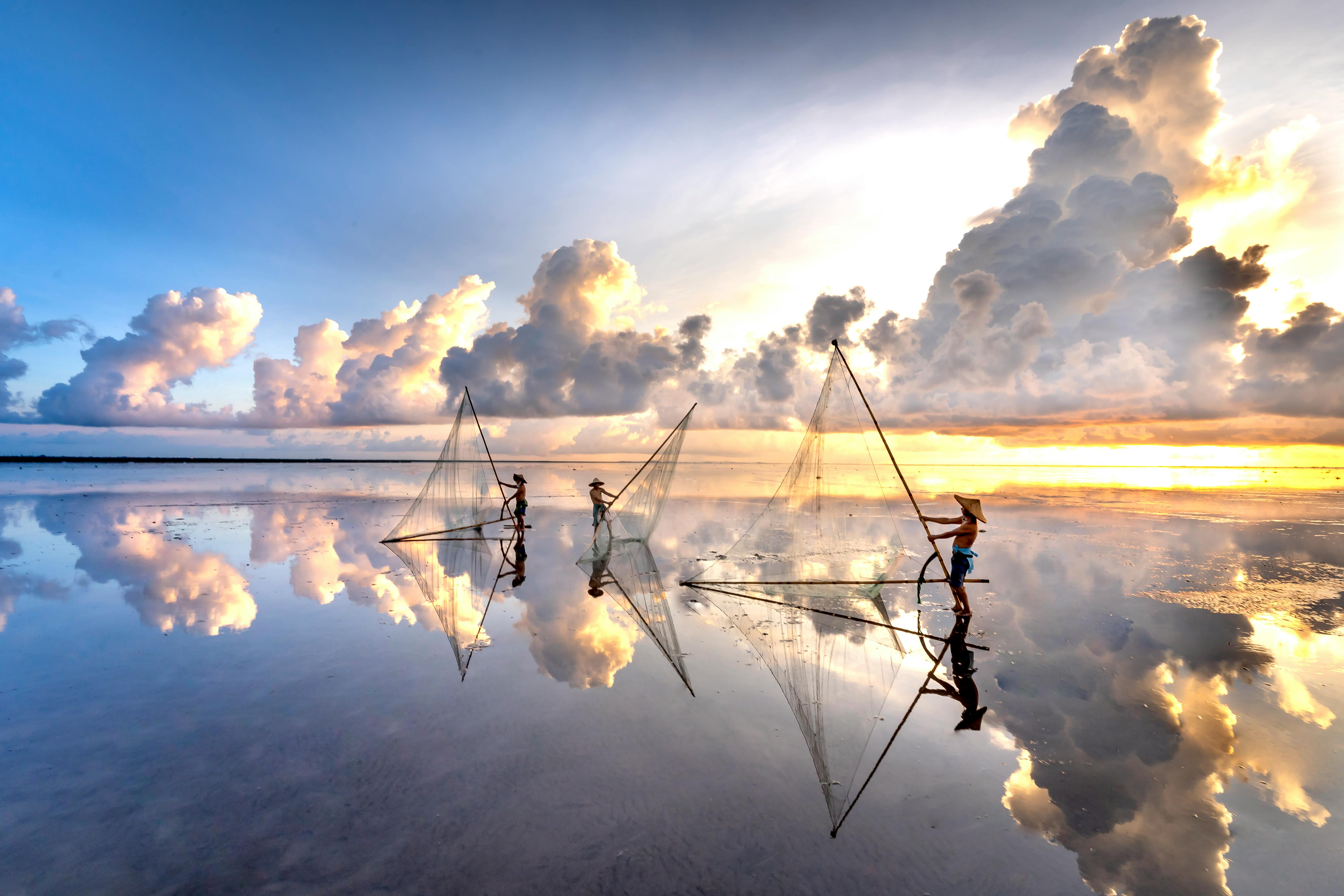Three fishermen casting nets, reflected on calm waters under a dramatic sunrise sky with clouds.