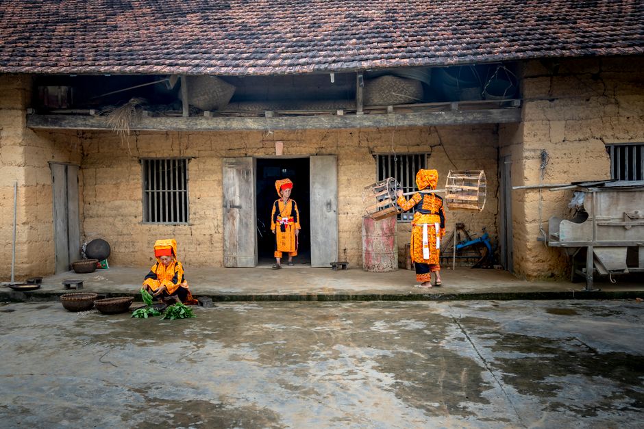 A respectful slow-travel moment in an ethnic village in Quang Ninh, Vietnam, showing daily life activities like weaving, cooking, and children playing near wooden houses, travelers observing quietly, soft natural daylight, culturally sensitive, highly detailed